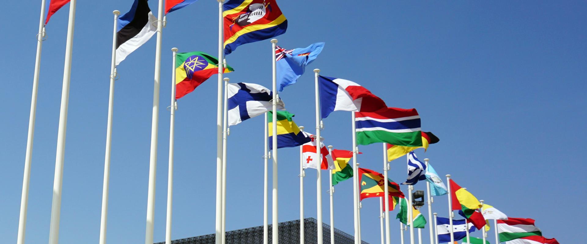 Multiple national flags hanging on flag poles against blue sky with a highrise building in the background